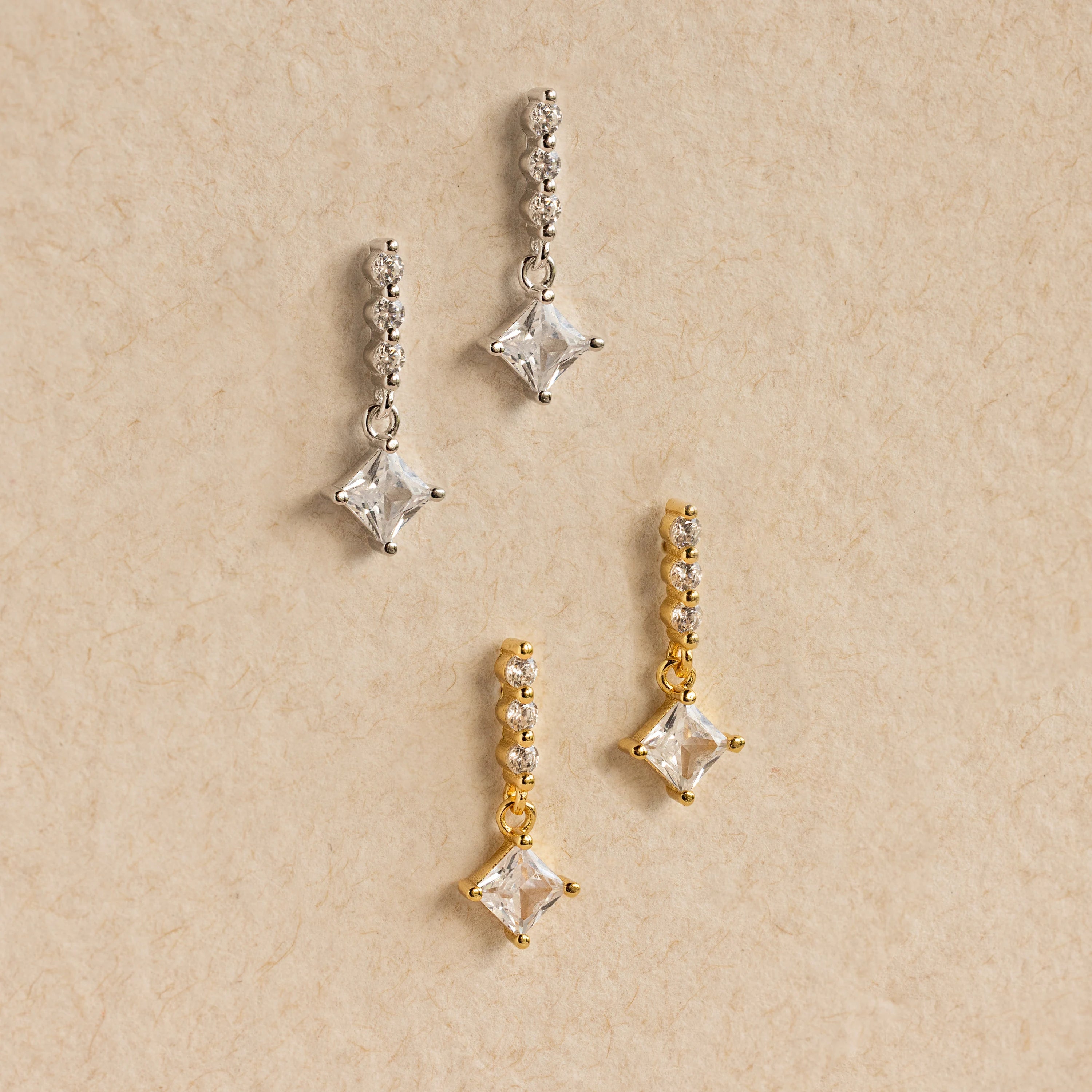 Two pairs of Princess Diamond Drop Studs—one in silver, one in gold—adorn a beige textured background, each pair featuring modern square crystals that highlight the sparkle of a princess cut gemstone.