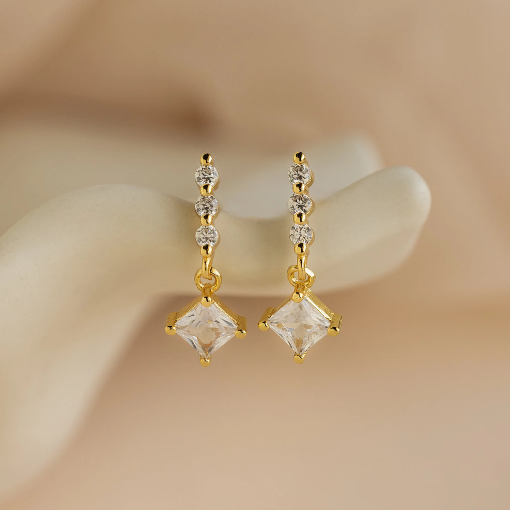 Princess Diamond Drop Studs featuring square princess-cut gemstone pendants and three small round gems, displayed on a cream background.