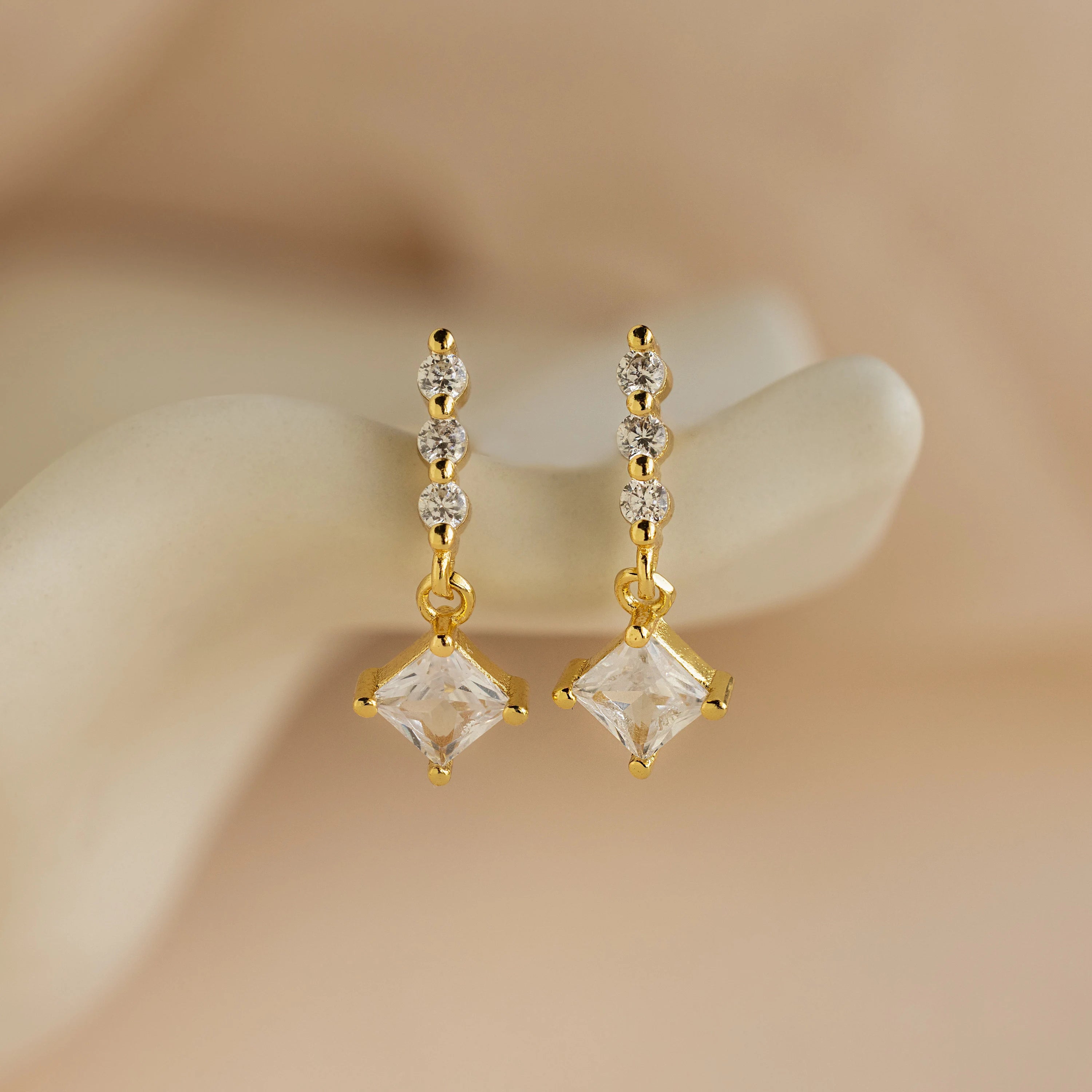 Princess Diamond Drop Studs featuring square princess-cut gemstone pendants and three small round gems, displayed on a cream background.
