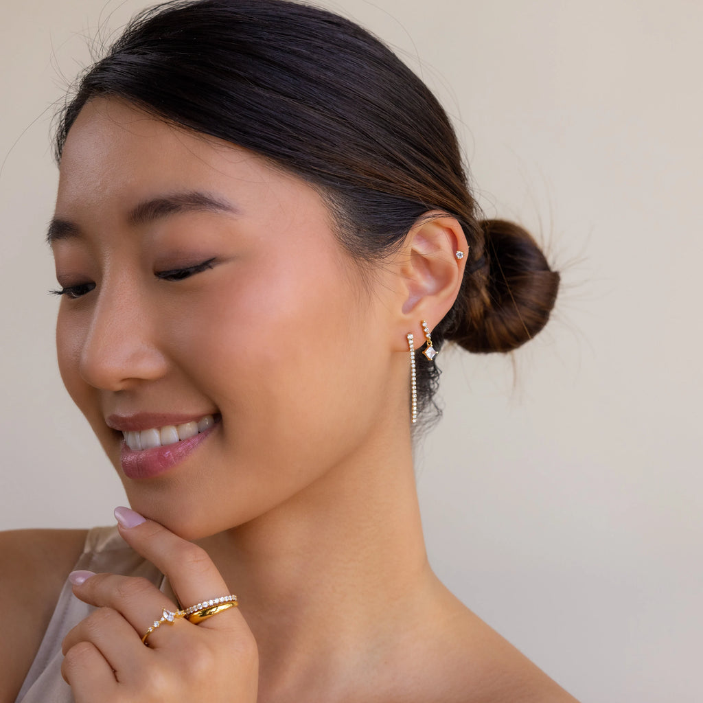Smiling woman with a low bun, wearing Princess Diamond Drop Studs and gold rings, looking down.