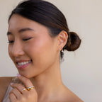 Smiling woman with a low bun, wearing Princess Diamond Drop Studs and gold rings, looking down.