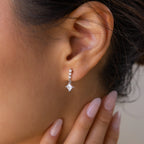 Close-up of a woman's ear adorned with Princess Diamond Drop Studs featuring a princess cut gemstone, her manicured hand resting gracefully near her face.