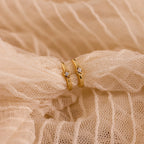 The Ericka Diamond Hoops—gold rings with small diamonds in a minimalist huggie style—are displayed on textured, cream-colored fabric.