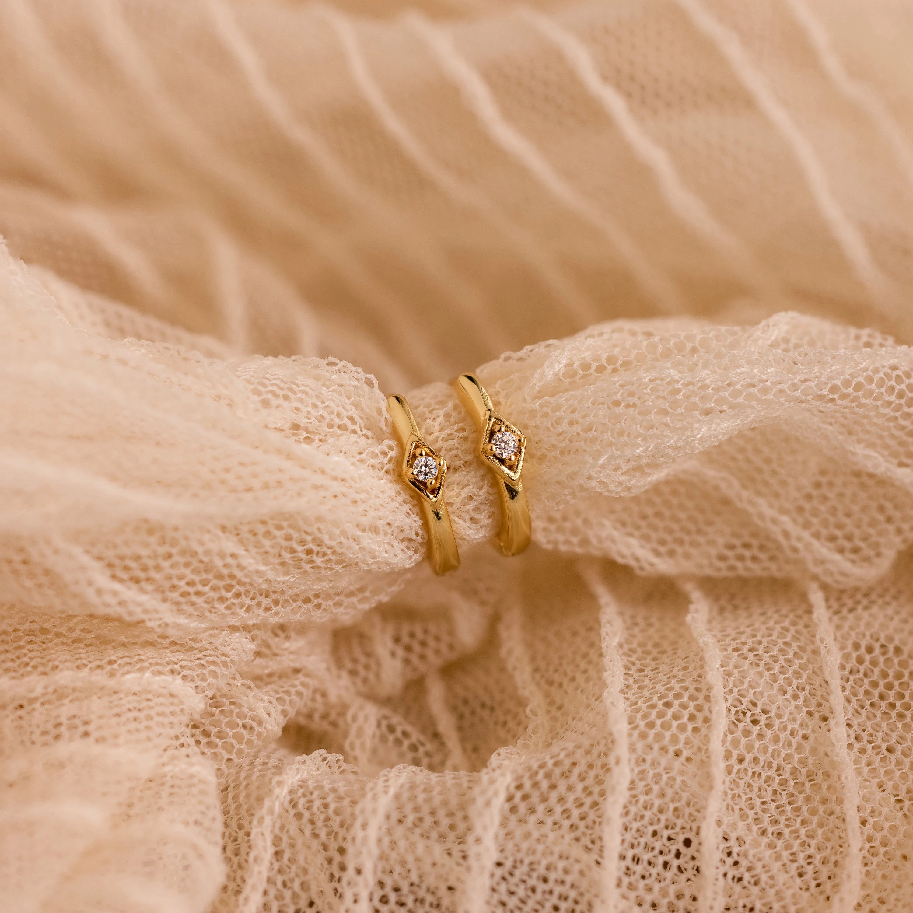 The Ericka Diamond Hoops—gold rings with small diamonds in a minimalist huggie style—are displayed on textured, cream-colored fabric.