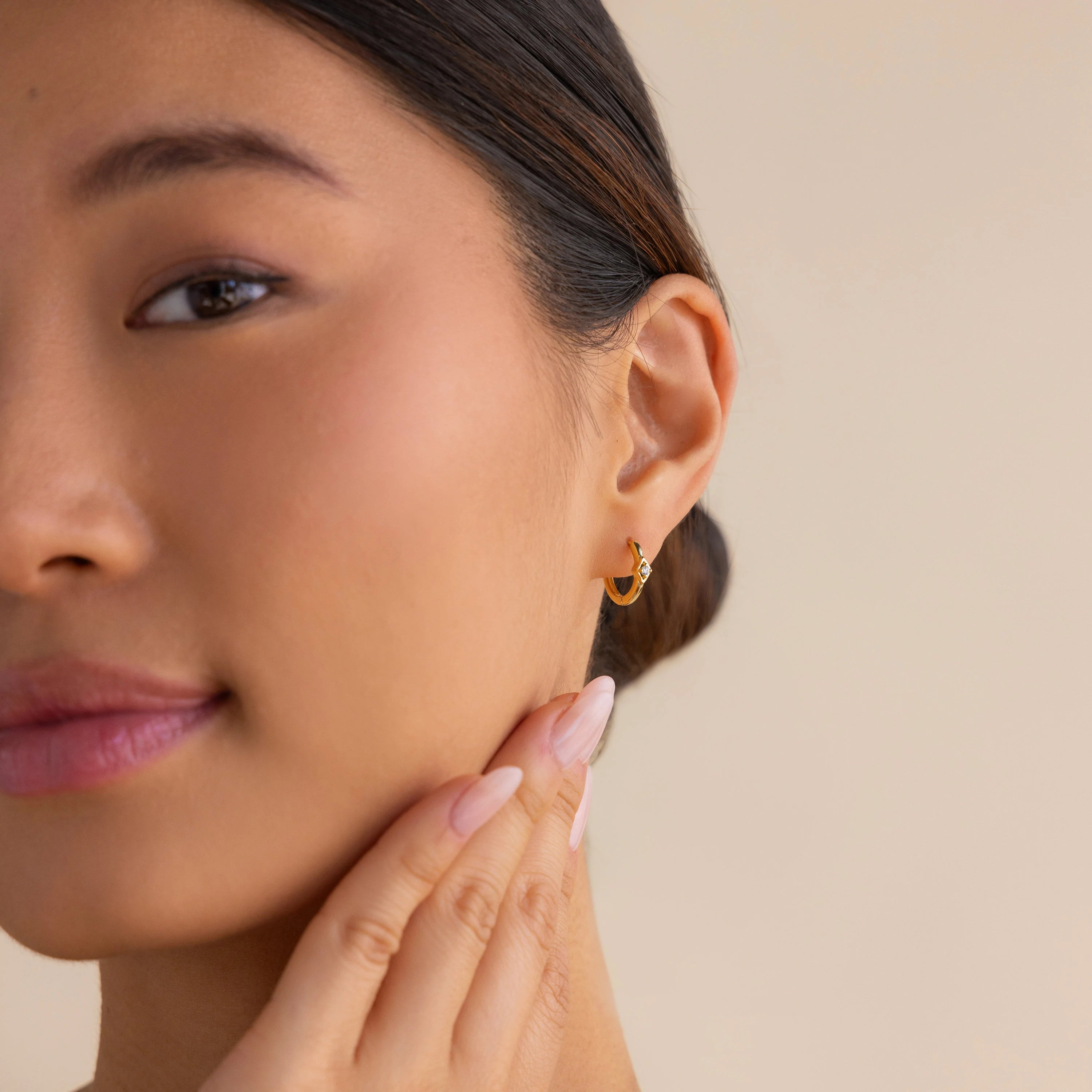 A woman with smooth skin touches her face, wearing Ericka Diamond Hoops and nude nail polish, her hair tied back.