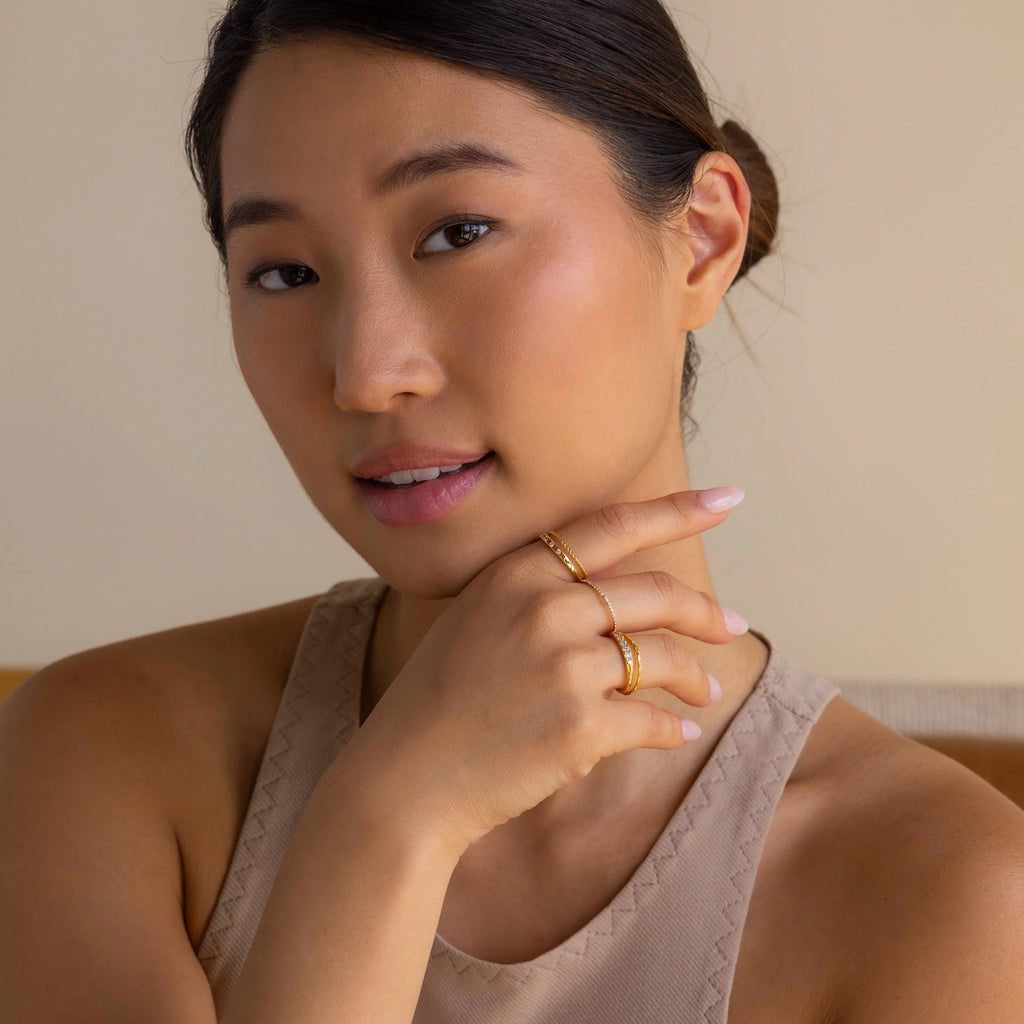 A woman with smooth skin, wearing the Diamond Station Ring and gold rings, poses in a beige top with her hand on her chin, gazing at the camera.