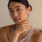 A woman with smooth skin, wearing the Diamond Station Ring and gold rings, poses in a beige top with her hand on her chin, gazing at the camera.