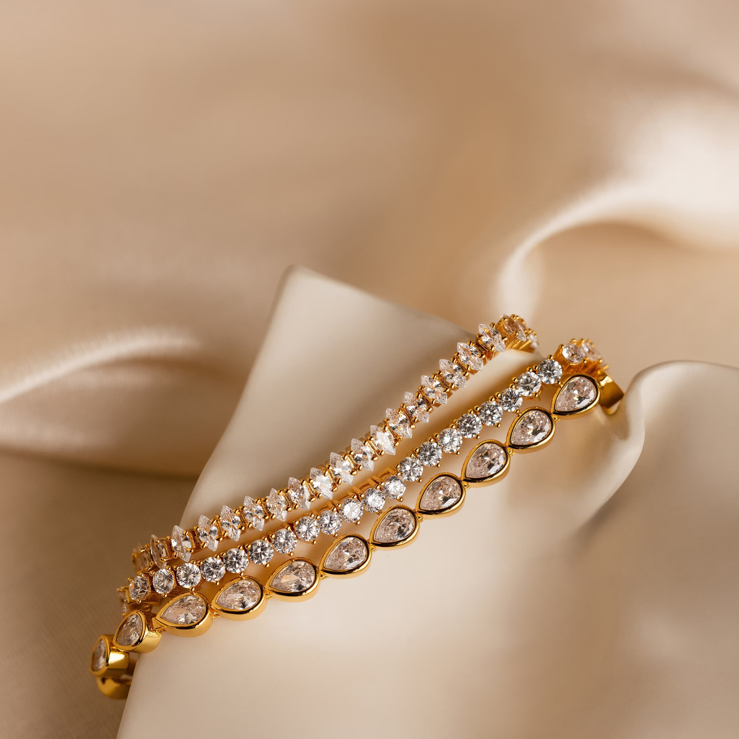The Pear Diamond Tennis Bracelet, crafted with pear-cut diamonds, is displayed on soft beige satin alongside two other gold and diamond bracelets.
