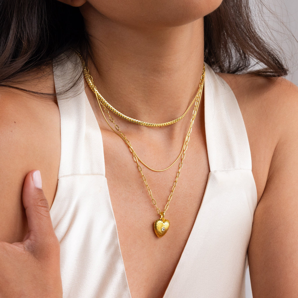A woman wears the Diamond Ribbed Heart Necklace layered with gold necklaces, paired with a cream sleeveless V-neck top.