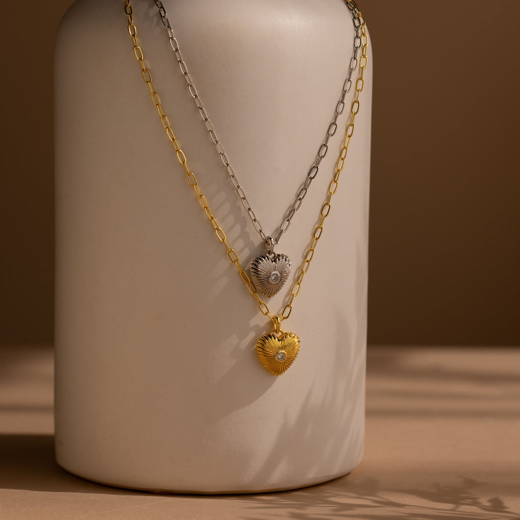 Two Diamond Ribbed Heart Necklaces, one in silver and one in gold, hang on chains draped over a neutral ceramic vase.