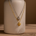 Two Diamond Ribbed Heart Necklaces, one in silver and one in gold, hang on chains draped over a neutral ceramic vase.