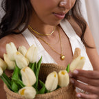 A woman in a white top holds a basket of white tulips and wears gold layered necklaces, including the Diamond Ribbed Heart Necklace, for a touch of vintage-inspired jewelry charm.