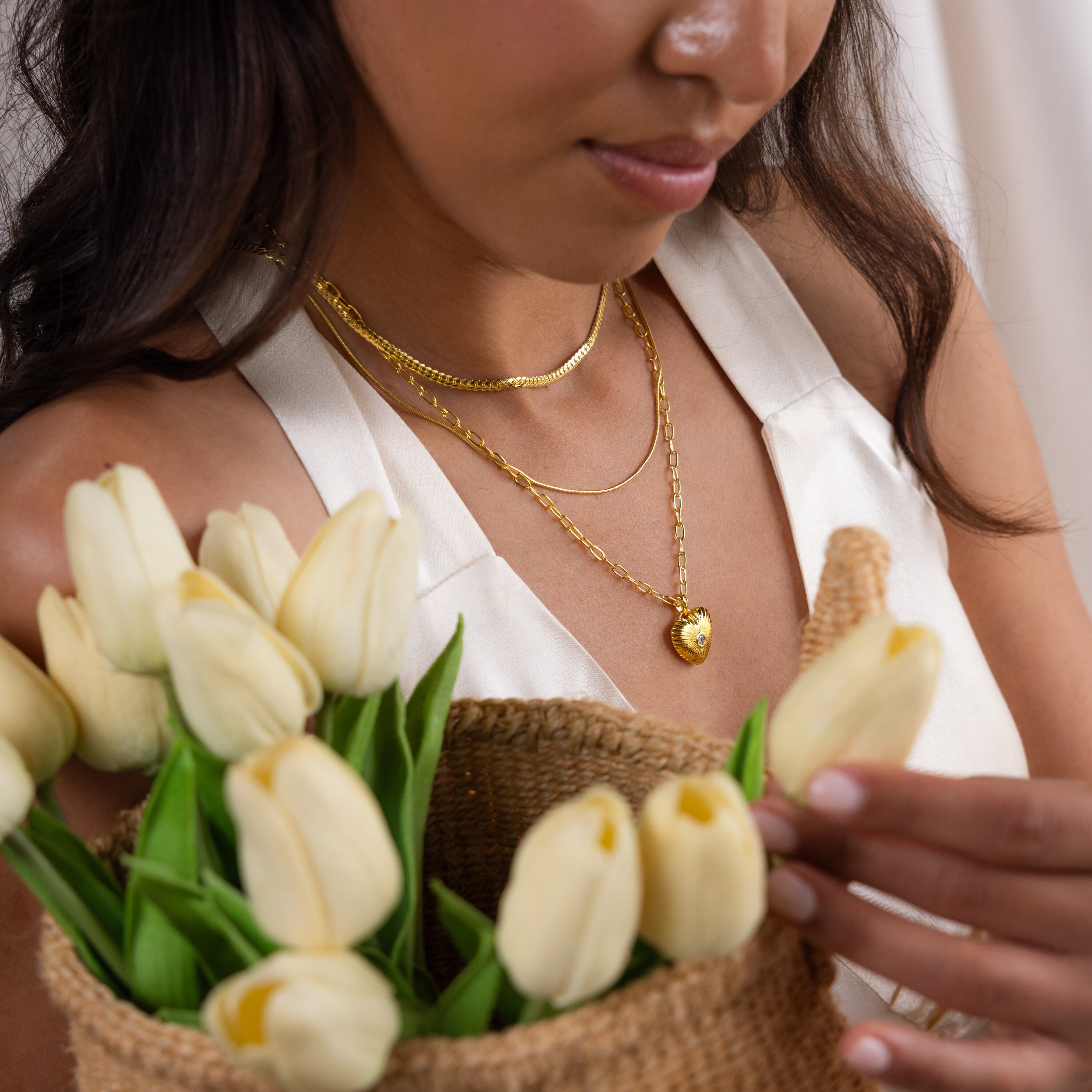 A woman in a white top holds a basket of white tulips and wears gold layered necklaces, including the Diamond Ribbed Heart Necklace, for a touch of vintage-inspired jewelry charm.