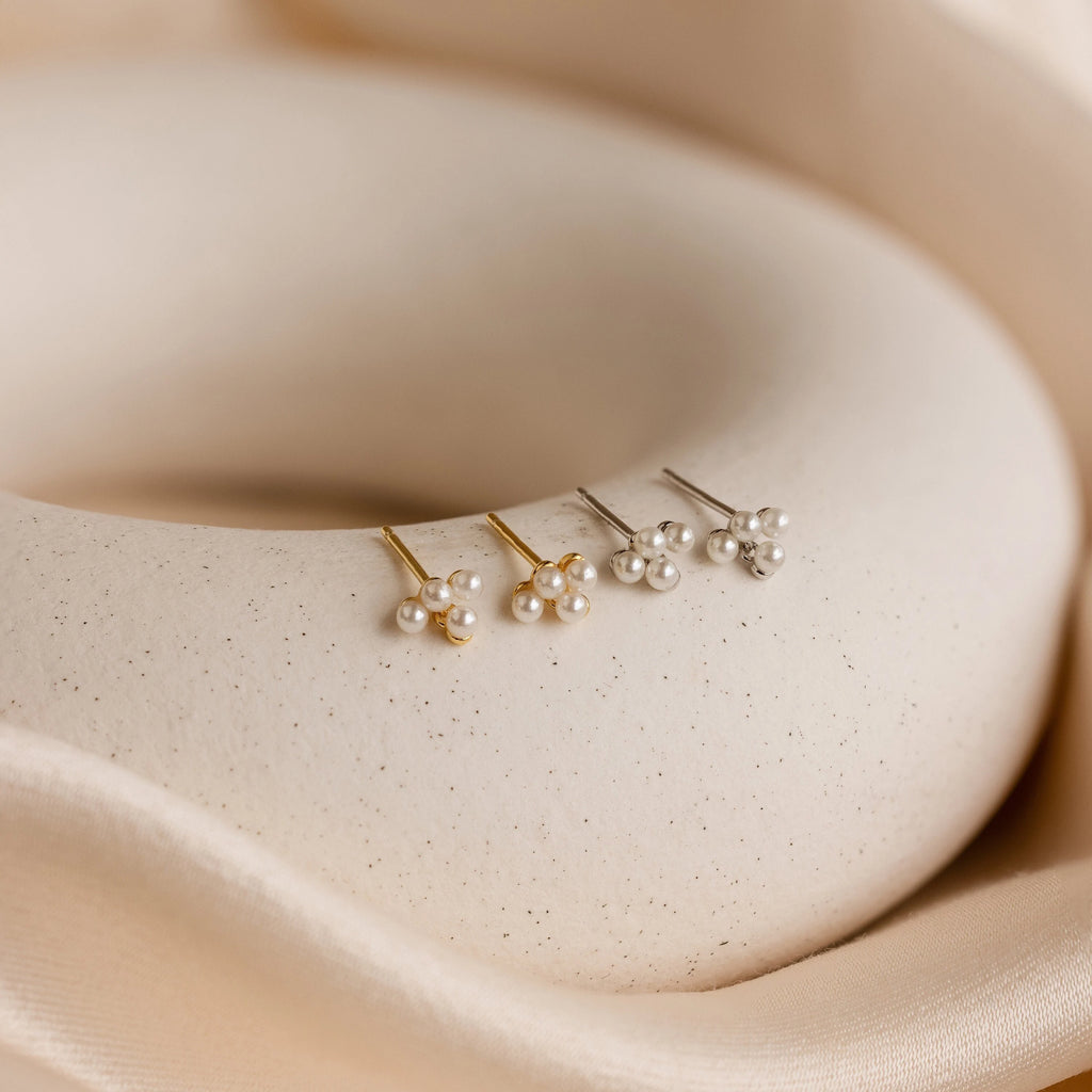 Three pairs of Pearl Cluster Drop Studs—two silver, one gold—rest on a smooth, cream surface with fabric folds, making them the perfect bridal earrings or a sophisticated wedding accessory.