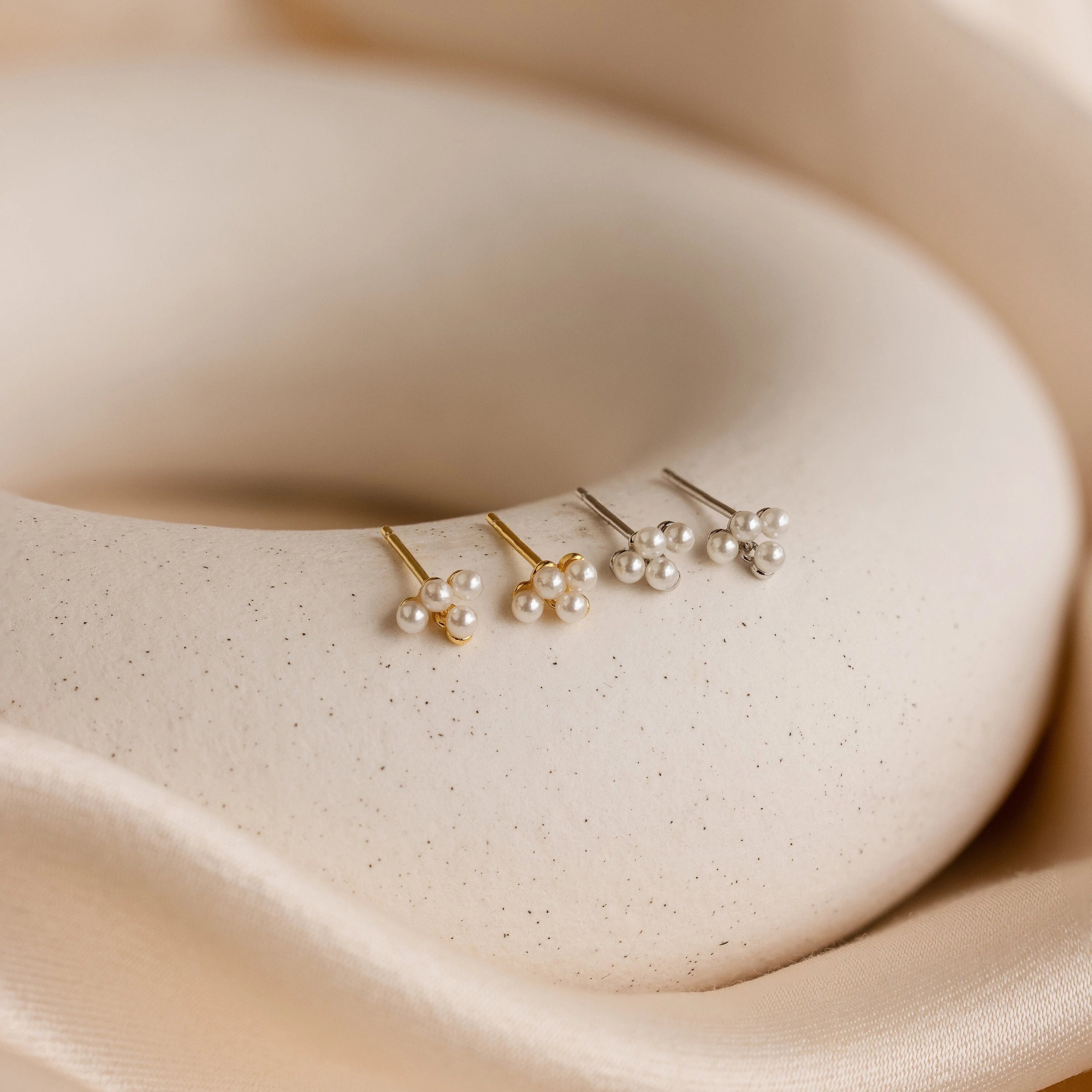 Three pairs of Pearl Cluster Drop Studs—two silver, one gold—rest on a smooth, cream surface with fabric folds, making them the perfect bridal earrings or a sophisticated wedding accessory.