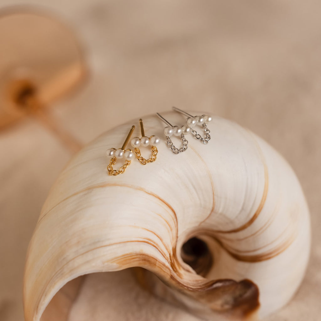 Two pairs of Pearl Bar Chain Studs, in gold and silver, are beautifully displayed on a large seashell.
