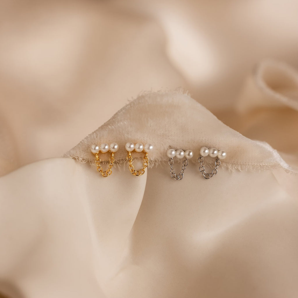 Two pairs of Pearl Bar Chain Studs—one gold, one silver—rest on soft beige fabric. These coquette earrings offer a timeless look with their elegant pearl accents and sleek bar chain design.