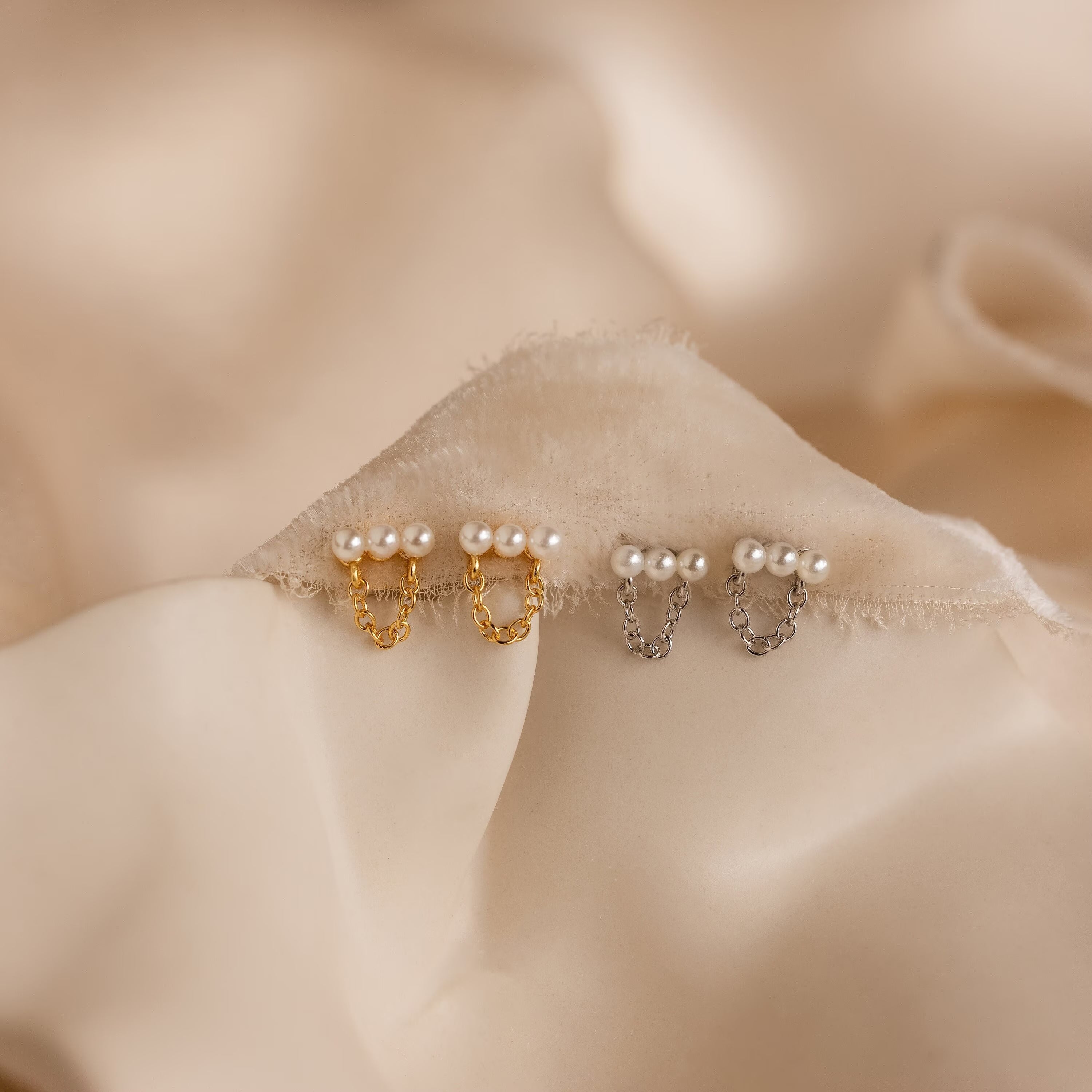 Two pairs of Pearl Bar Chain Studs—one gold, one silver—rest on soft beige fabric. These coquette earrings offer a timeless look with their elegant pearl accents and sleek bar chain design.