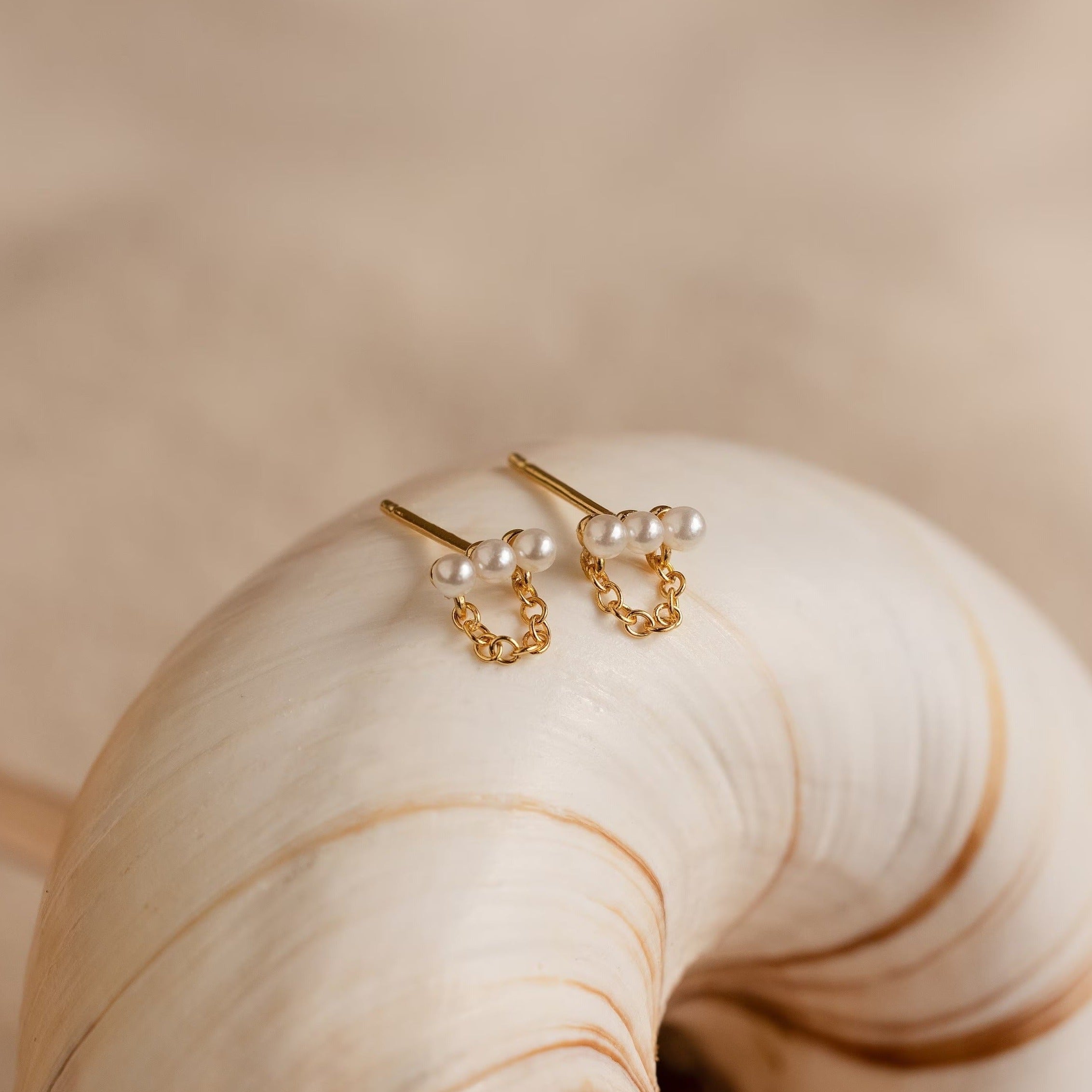 The Pearl Bar Chain Studs, featuring small pearls and delicate chain details, are elegantly displayed on a large white seashell.
