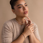 A woman gazes thoughtfully to the side against a beige background, wearing gold jewelry and a sparkling 3 Prong Tennis Bracelet with her cozy beige sweater.