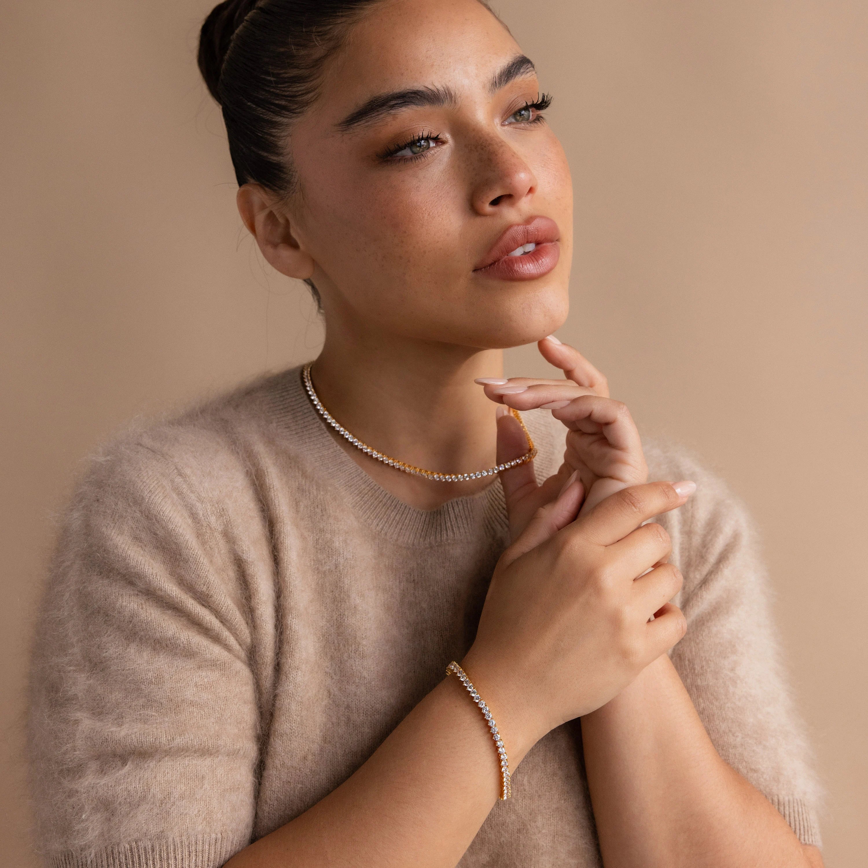 A woman gazes thoughtfully to the side against a beige background, wearing gold jewelry and a sparkling 3 Prong Tennis Bracelet with her cozy beige sweater.