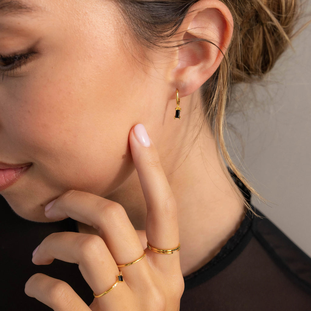 A woman wearing gold rings and Onyx Baguette Drop Huggies touches her face, highlighting the elegance of her Onyx Baguette Drop Huggies earrings.