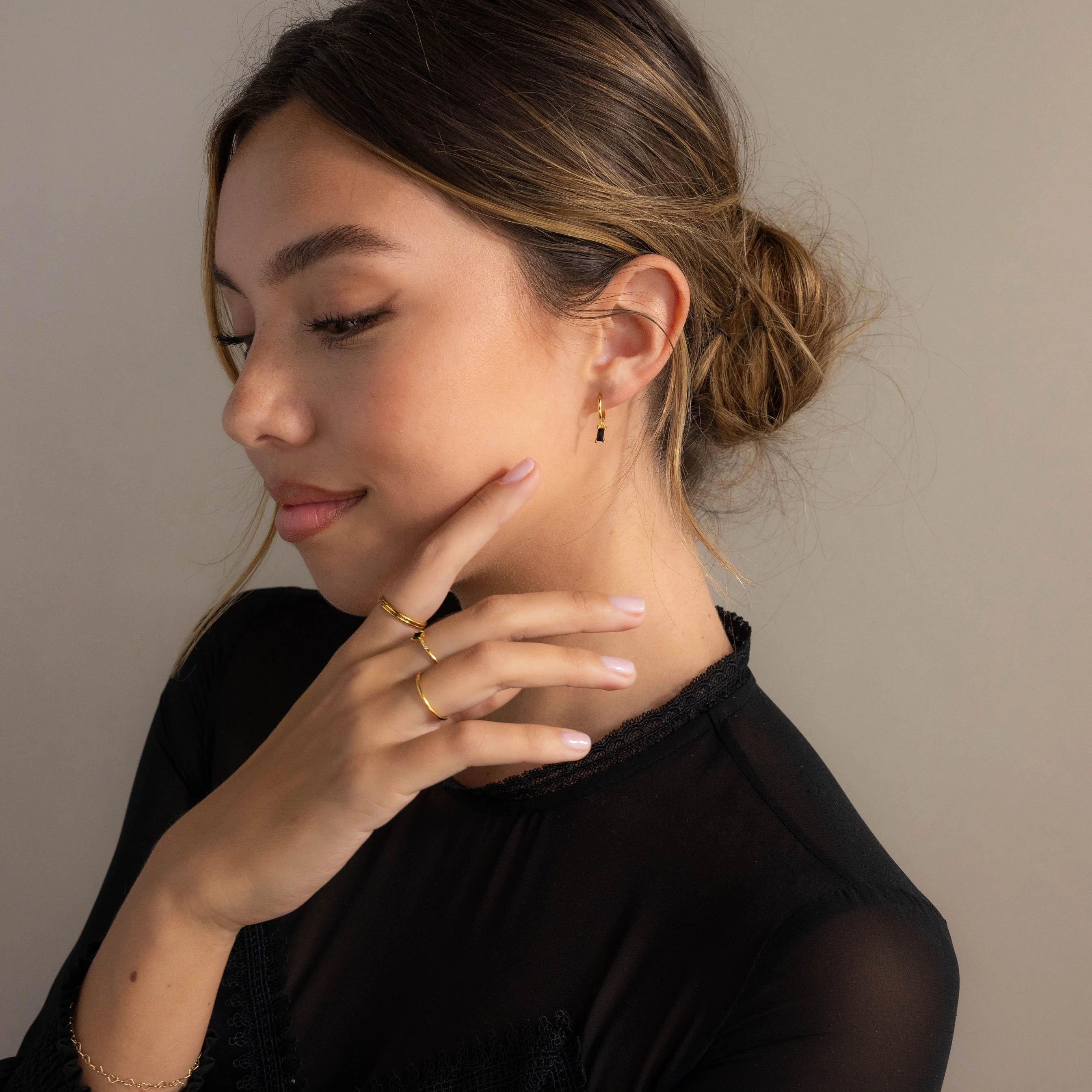 A woman with brown hair in a low bun, wearing gold jewelry and the Onyx Baguette Drop Huggies, dressed in a black top, gazes to the side with a soft smile.