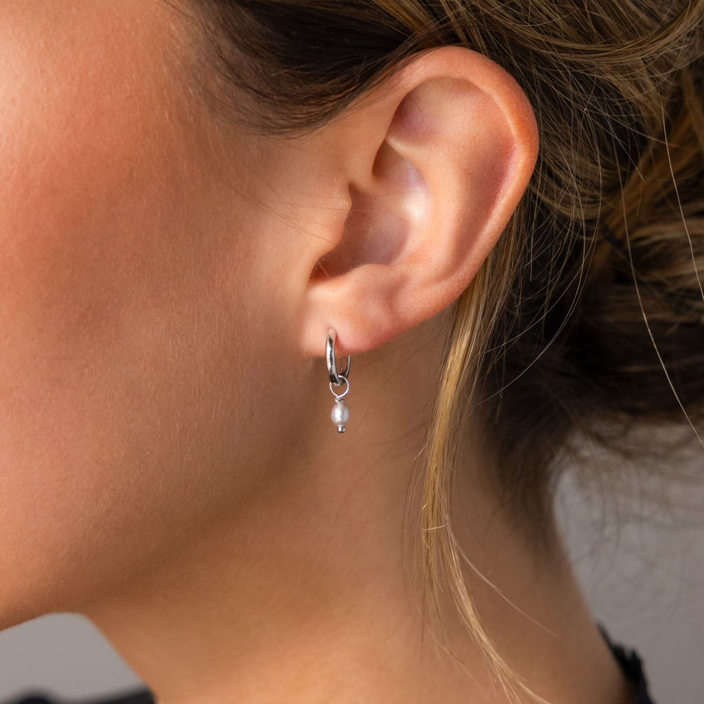 Close-up of a woman's ear wearing the Smokey Pearl Drop Huggies, small silver hoops featuring a delicate white pearl charm.
