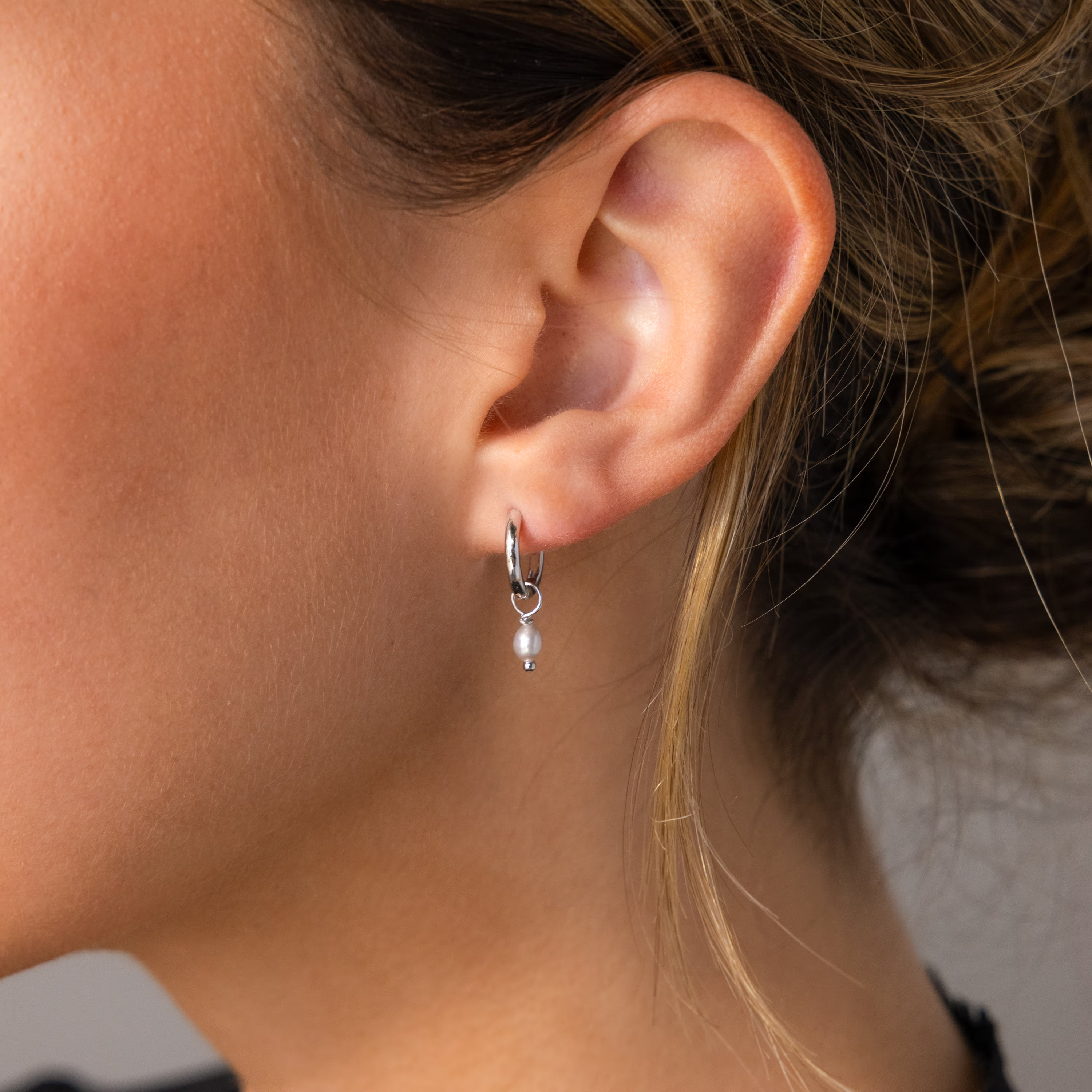 Close-up of a woman's ear wearing the Smokey Pearl Drop Huggies, small silver hoops featuring a delicate white pearl charm.