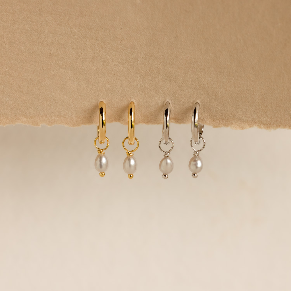 Two pairs of Smokey Pearl Drop Huggies—one gold, one silver—hang on textured paper. These elegant huggie hoop earrings feature delicate pearl drops to add a charming touch to any outfit.