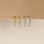 Two pairs of Smokey Pearl Drop Huggies—one gold, one silver—hang on textured paper. These elegant huggie hoop earrings feature delicate pearl drops to add a charming touch to any outfit.