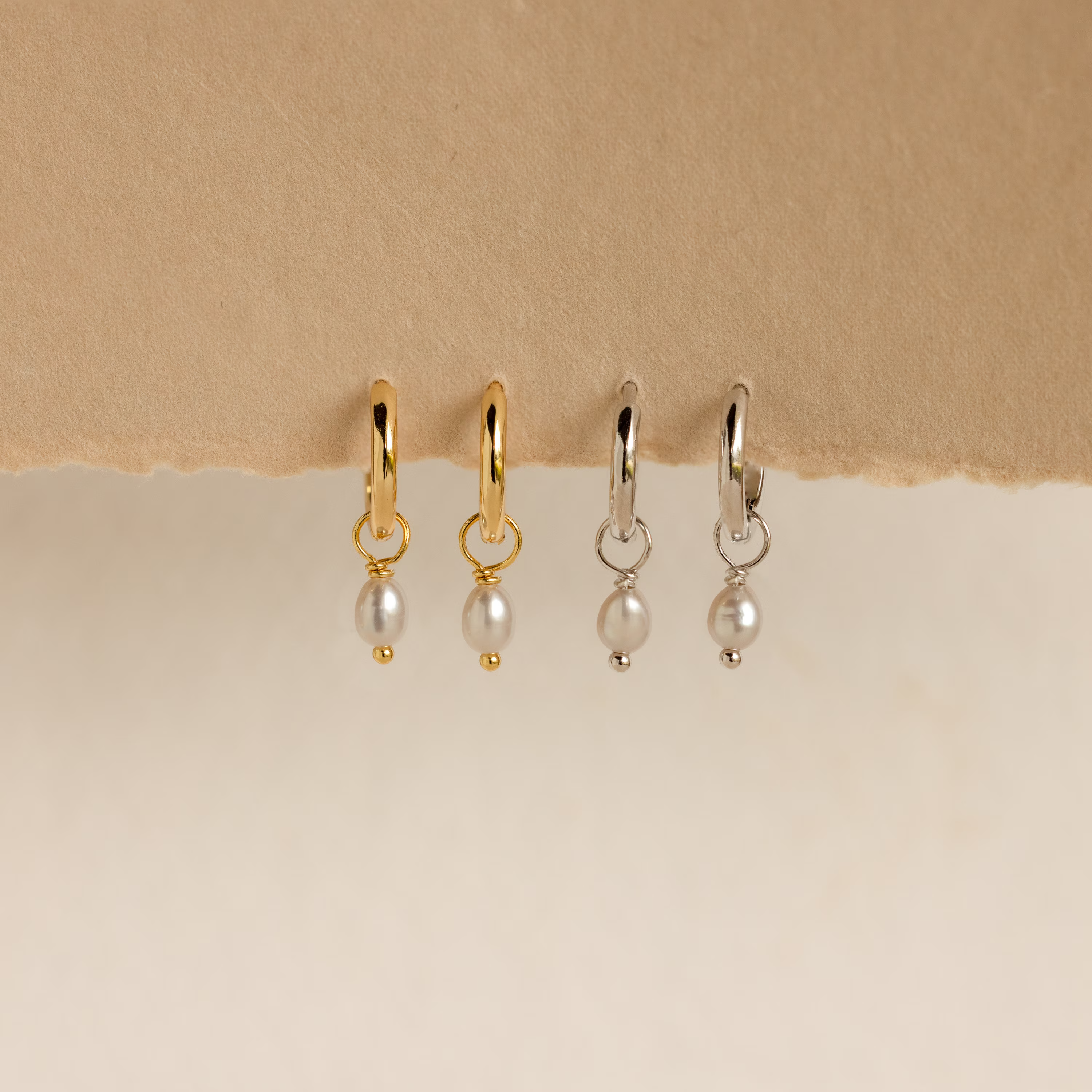 Two pairs of Smokey Pearl Drop Huggies—one gold, one silver—hang on textured paper. These elegant huggie hoop earrings feature delicate pearl drops to add a charming touch to any outfit.