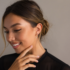 A woman with a low bun smiles, wearing a black top and Smokey Pearl Drop Huggies, gently touching her chin as the pearl charm on the earrings catches the light.