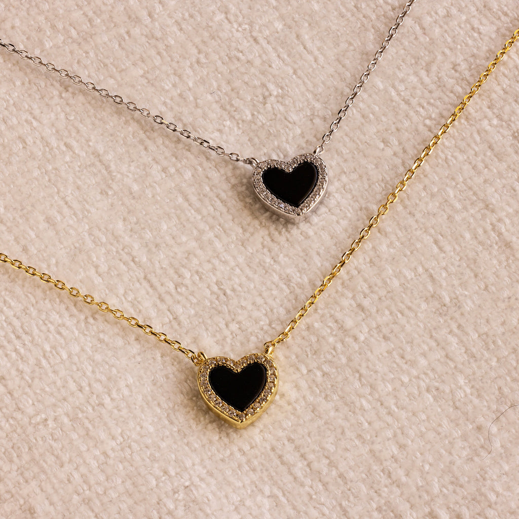 The Pave Onyx Heart Necklace, available with either a gold or silver chain, rests on a textured beige surface, showcasing an elegant heart-shaped charm design.