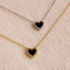 The Pave Onyx Heart Necklace, available with either a gold or silver chain, rests on a textured beige surface, showcasing an elegant heart-shaped charm design.