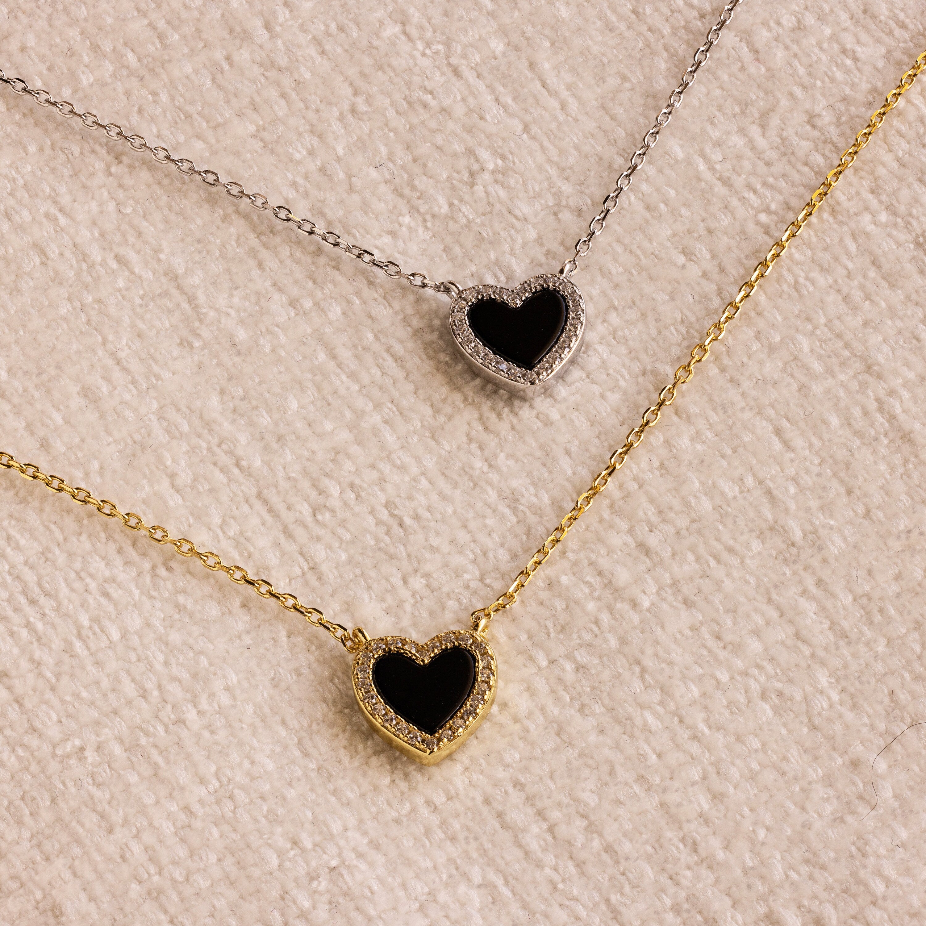 The Pave Onyx Heart Necklace, available with either a gold or silver chain, rests on a textured beige surface, showcasing an elegant heart-shaped charm design.