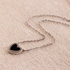 The Pave Onyx Heart Necklace in Sterling Silver features an onyx heart pendant outlined with sparkling pave CZ diamonds, displayed elegantly on a soft beige surface.