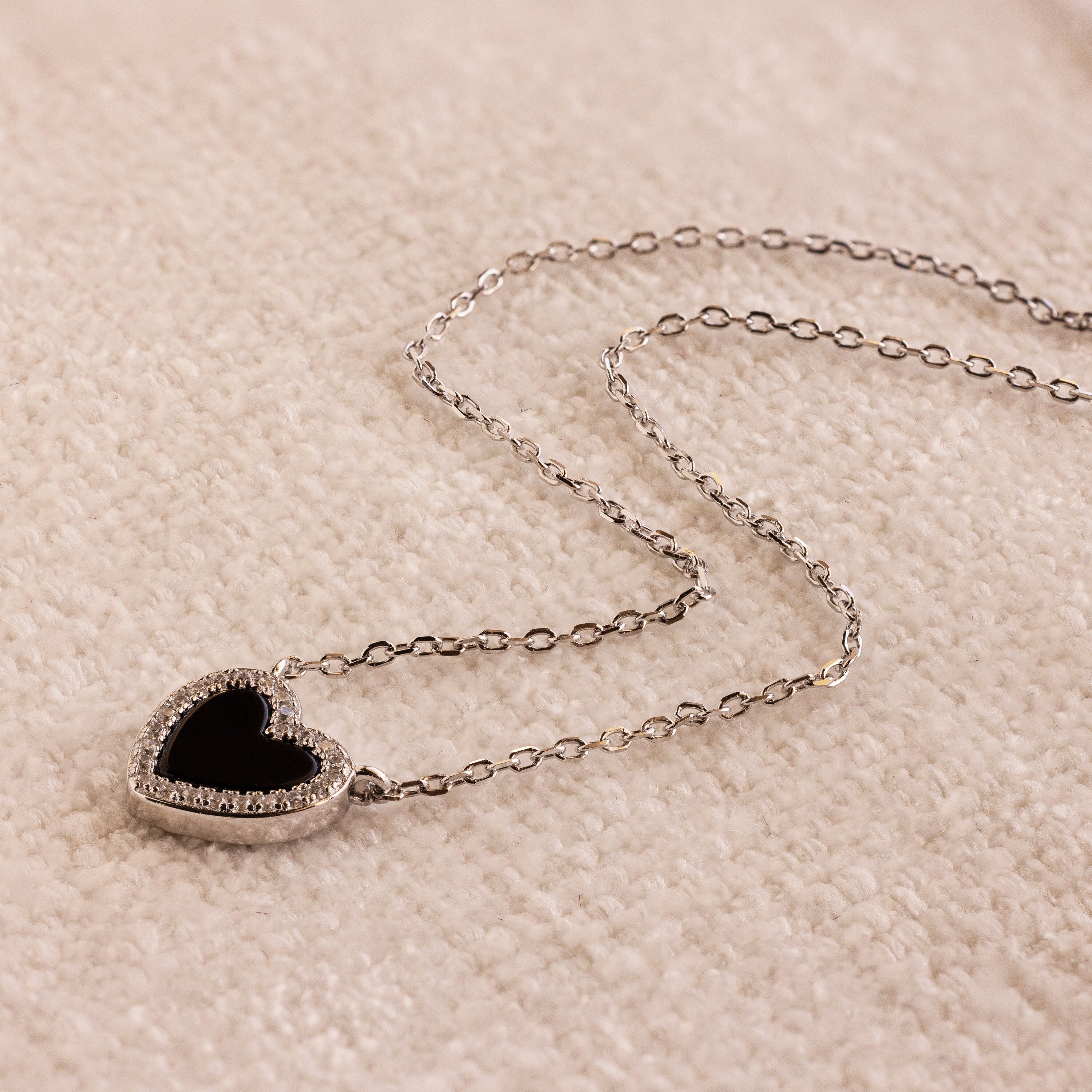 The Pave Onyx Heart Necklace in Sterling Silver features an onyx heart pendant outlined with sparkling pave CZ diamonds, displayed elegantly on a soft beige surface.