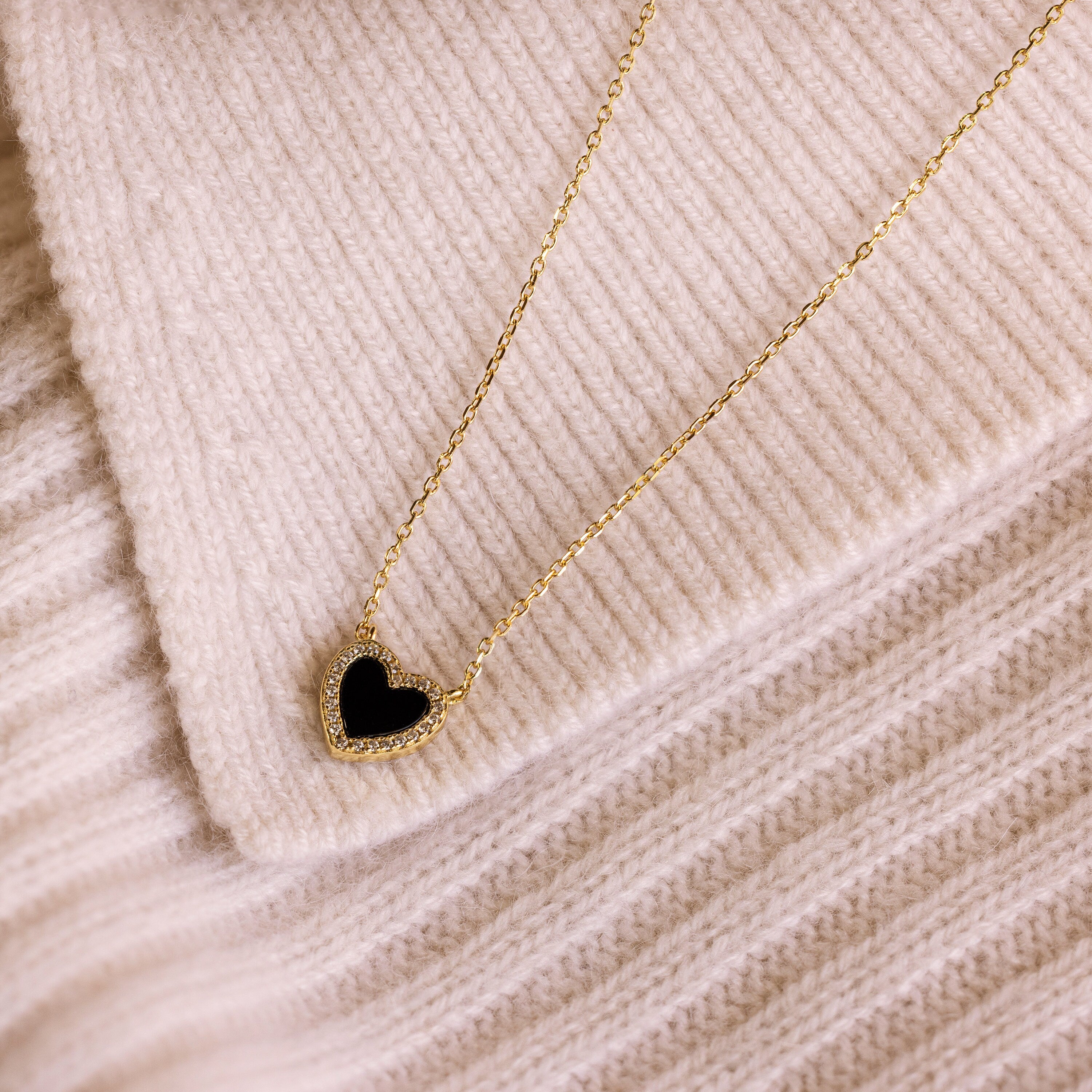 The Pave Onyx Heart Necklace in 18K Gold features a black heart-shaped charm pendant, elegantly displayed on cream-colored ribbed knit fabric.