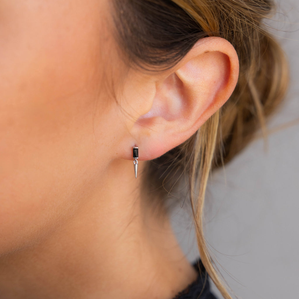 Close-up of a woman's ear wearing the Onyx Dagger Drop Earrings, featuring a small silver design with a baguette-cut onyx and pointed dagger charm.