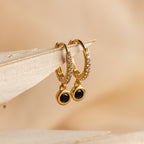 The Pave Onyx Drop Huggies showcase sparkling pavé stones and dangling black Onyx charms, beautifully displayed on a beige surface.