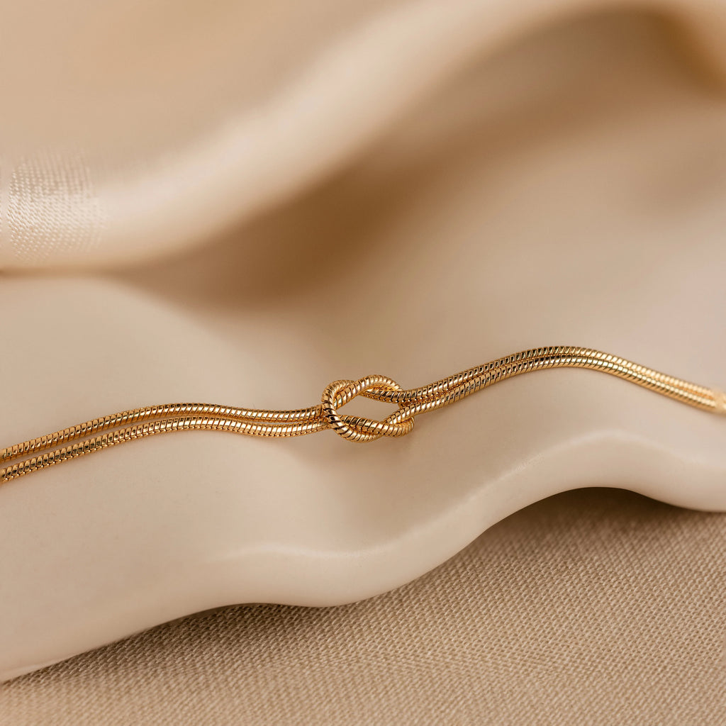 The Love Knot Bracelet features a delicate knot design with double chain detail, elegantly displayed on beige fabric—a perfect gift for couples.