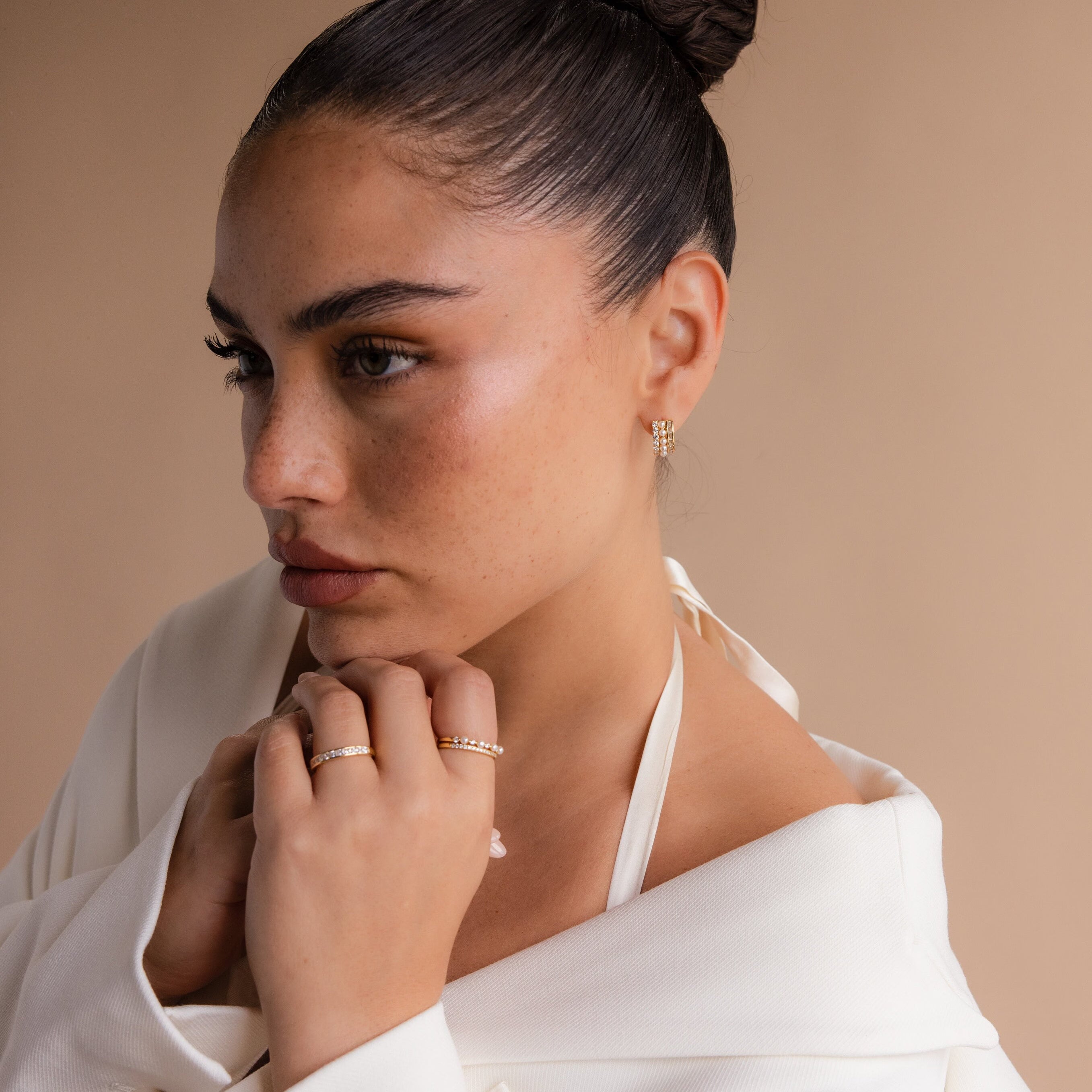A woman with a sleek bun wears Diamond Pearl Stacked Hoops bridal jewelry and a white top, gazing thoughtfully to the side.