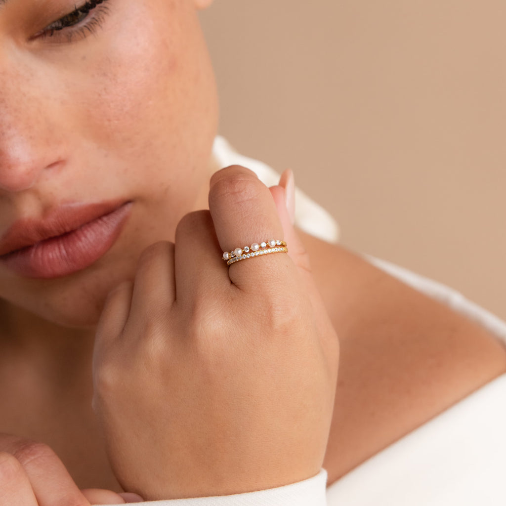 A close-up shows a woman's hand wearing the Pave Pearl Stacking Ring Set—two gold rings, including a delicate pave pearl design—held near her face against a beige background.