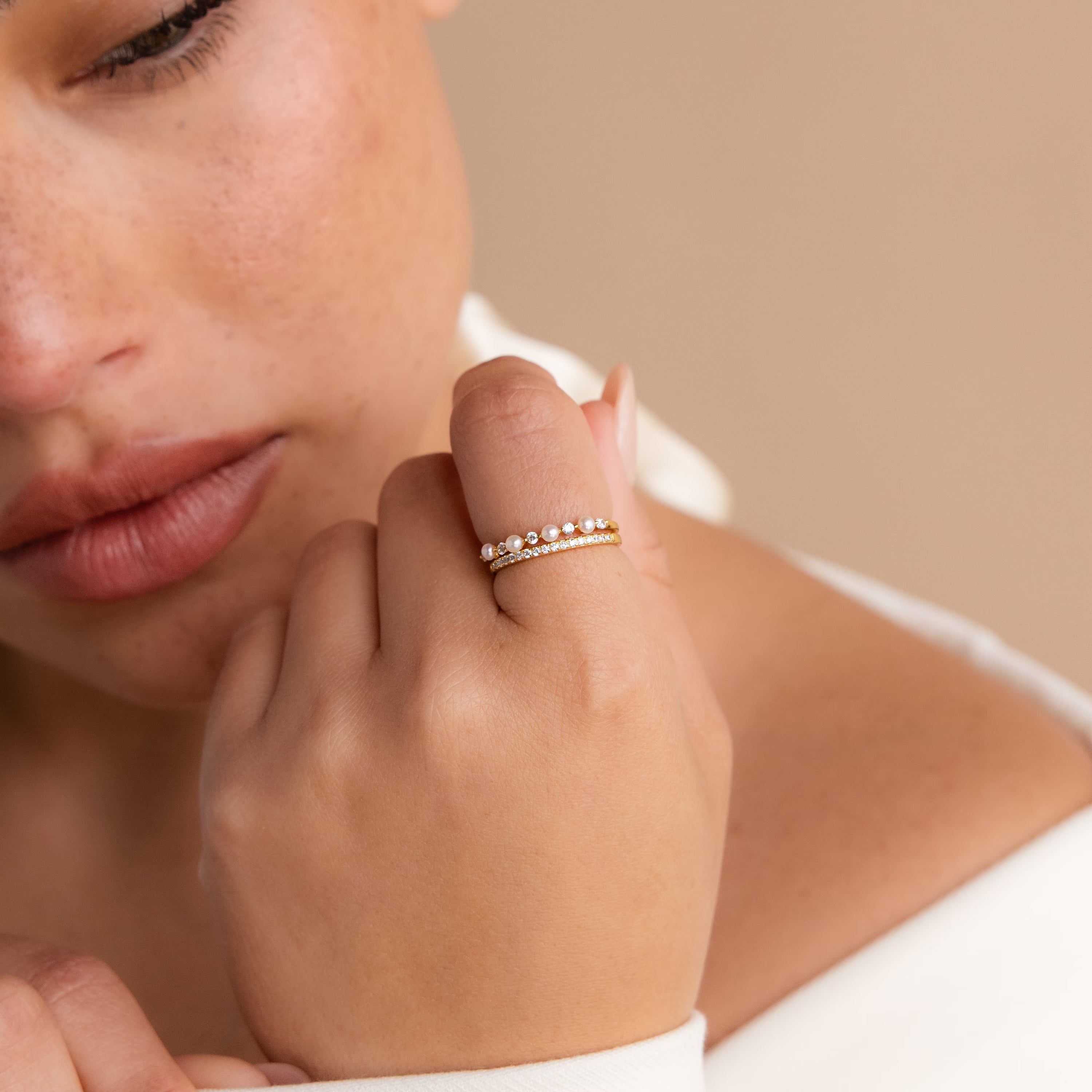 A close-up shows a woman's hand wearing the Pave Pearl Stacking Ring Set—two gold rings, including a delicate pave pearl design—held near her face against a beige background.
