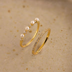 The Pave Pearl Stacking Ring Set features a gold pave pearl ring with small pearls and diamonds, plus a gold ring with a row of small diamonds, shown on a beige surface.