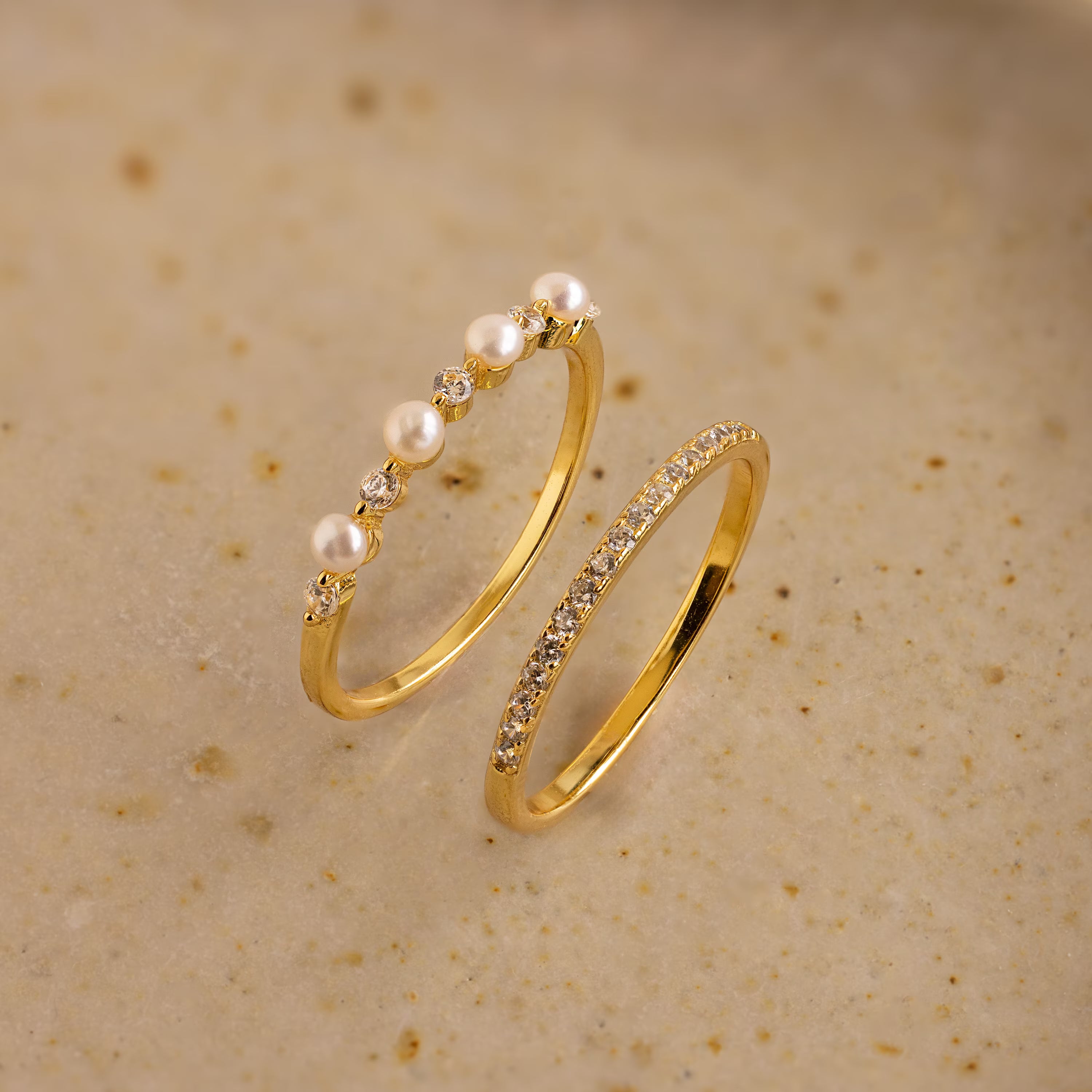 The Pave Pearl Stacking Ring Set features a gold pave pearl ring with small pearls and diamonds, plus a gold ring with a row of small diamonds, shown on a beige surface.
