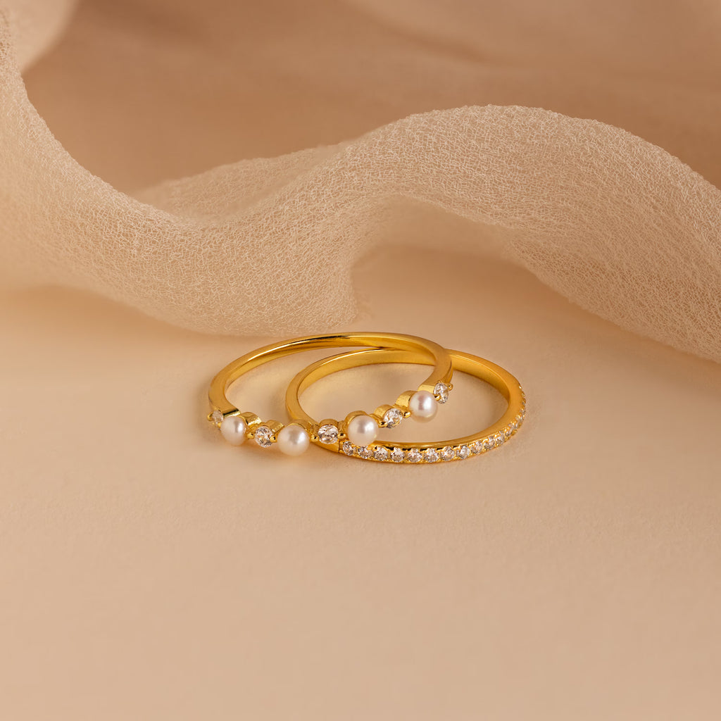 The Pave Pearl Stacking Ring Set, featuring elegant gold bands with shimmering small diamonds, is beautifully displayed on a beige fabric background, emphasizing its timeless charm and sophisticated design.