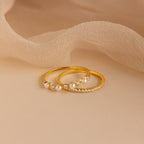 The Pave Pearl Stacking Ring Set, featuring elegant gold bands with shimmering small diamonds, is beautifully displayed on a beige fabric background, emphasizing its timeless charm and sophisticated design.