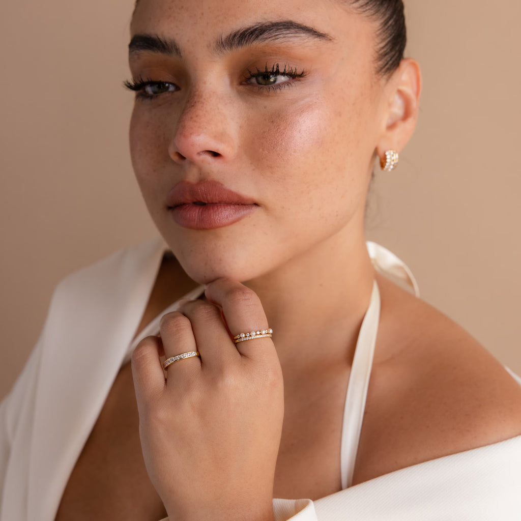 Woman with dewy skin gazing confidently, wearing a white halter top and the Pave Pearl Stacking Ring Set, along with gold gemstone rings and an earring.